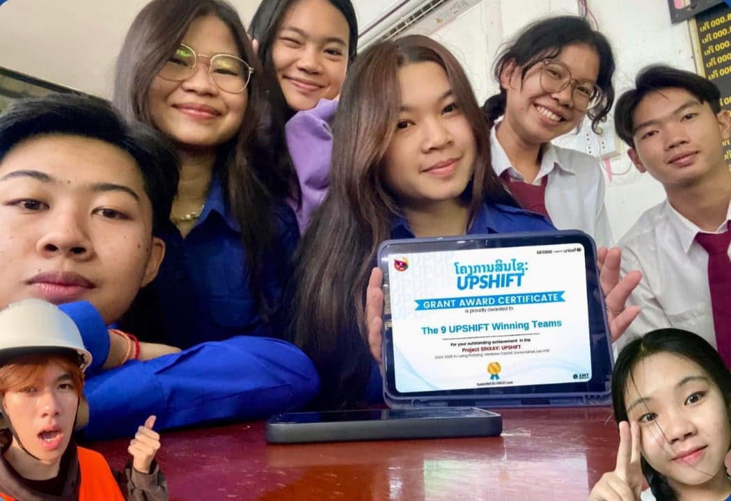Team BSF Initiative, the Black Soldier Fly project from Vientiane — eight participants in a close indoor group selfie, one in a safety helmet and vest, one holding a tablet displaying their grant award certificate.