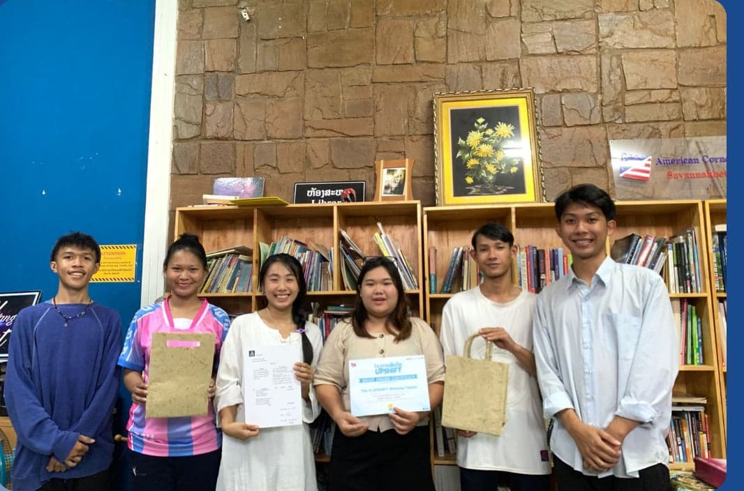 The Phoenix Fold team, the Eco Rice Straw project from Savannakhet — six participants standing indoors in front of a bookshelf, two holding eco-friendly paper bags made from rice straw and one holding their grant certificate.