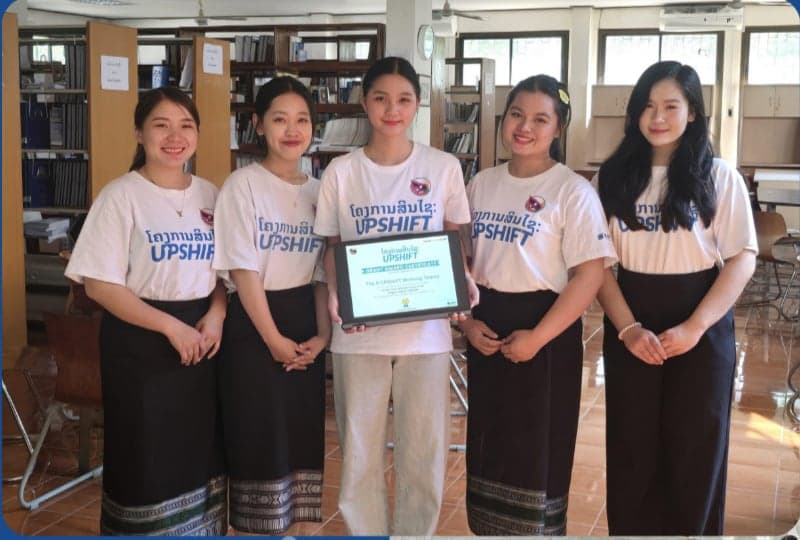 Team Dreamer, the Green Market project from Luang Prabang — five young women in white UPSHIFT shirts and traditional Lao sinh skirts, one holding their grant certificate, photographed in a library.
