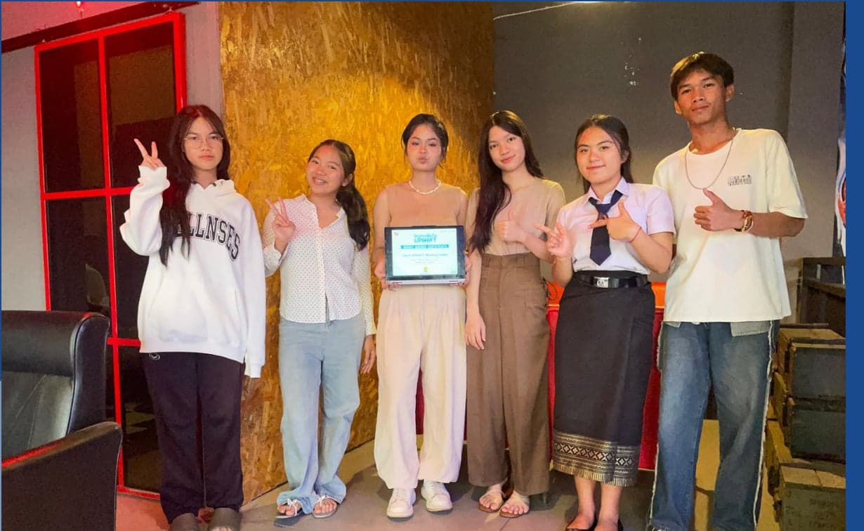 Team Luther, the No Waste Better Drain project from Savannakhet — six participants standing indoors making peace signs, one holding a tablet displaying their grant certificate.