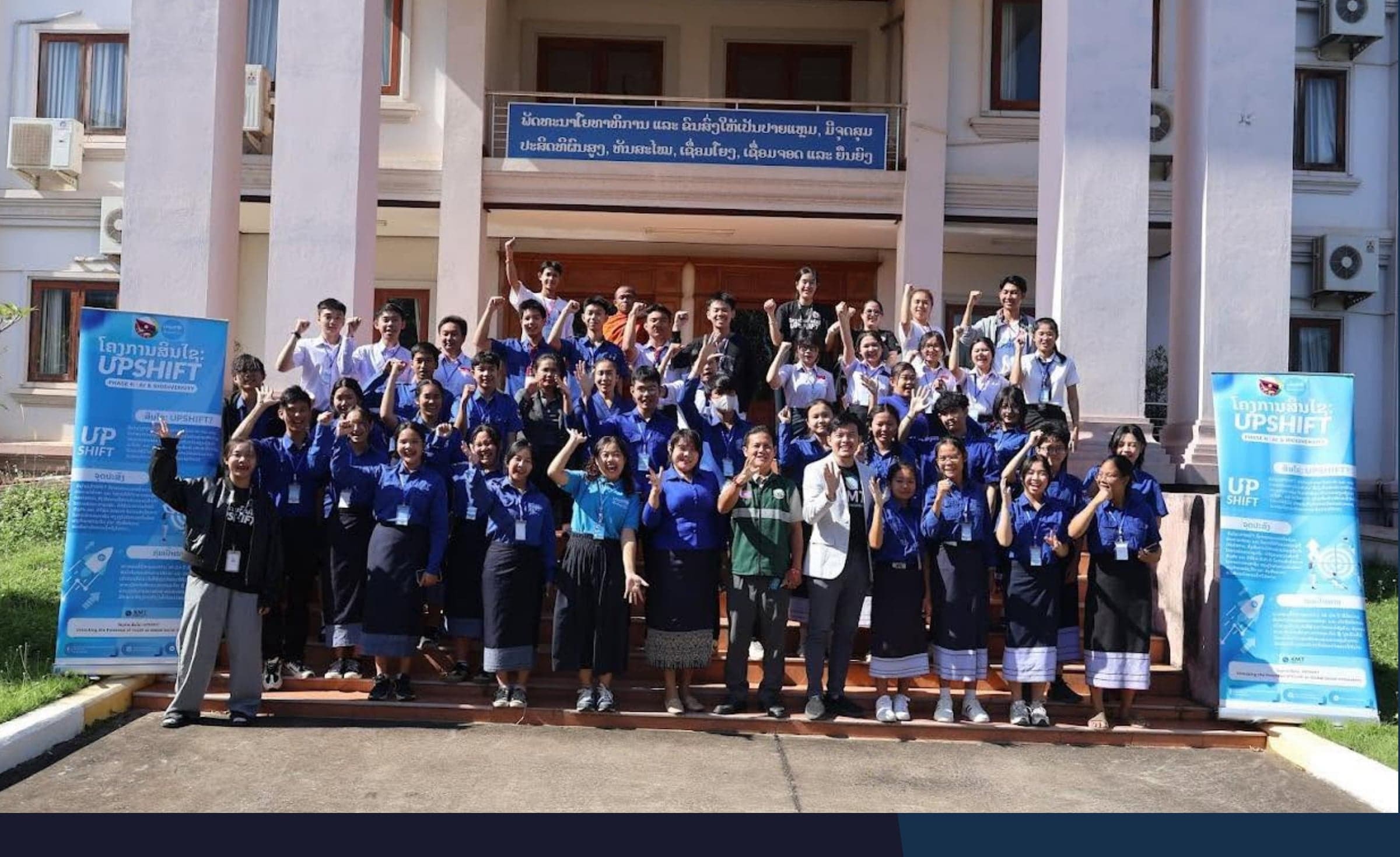 Sinxay UPSHIFT participants and staff celebrating together on the steps of a government building, many raising a fist in the air, with UPSHIFT roll-up banners on either side.